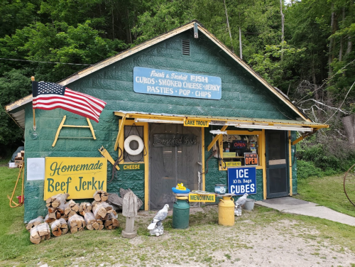 A rustic green building with signs for fish, jerky, and snacks, featuring an American flag and stacked firewood outside.