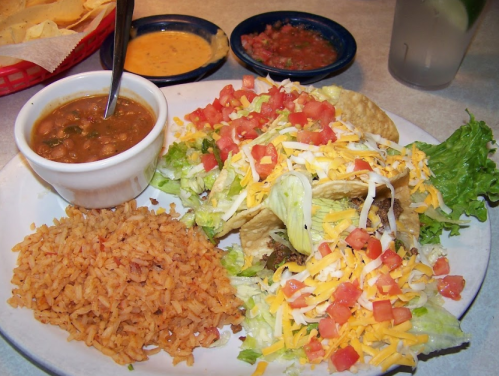 A plate of tacos topped with lettuce, tomatoes, and cheese, served with rice and beans, alongside salsa and cheese dip.