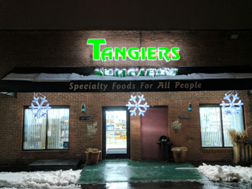 Storefront of "Tangiers" with neon green sign, snowflakes, and specialty food offerings, surrounded by snow.