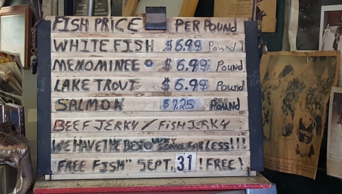 Sign displaying fish prices: White Fish $6.99, Menominee $6.99, Lake Trout $6.99, Salmon $7.25, Beef Jerky, Free Fish Sept 31.