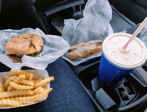 A car interior with a burger, hot dog, crinkle-cut fries, and a drink in a cup on the center console.