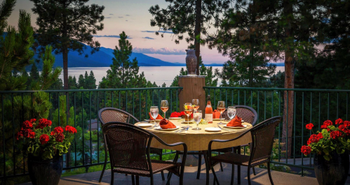 A beautifully set outdoor dining table with drinks and flowers, overlooking a serene lake and mountains at sunset.