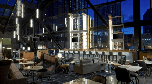 Modern bar interior with sleek furniture, large windows, and ambient lighting, set against a nighttime city backdrop.