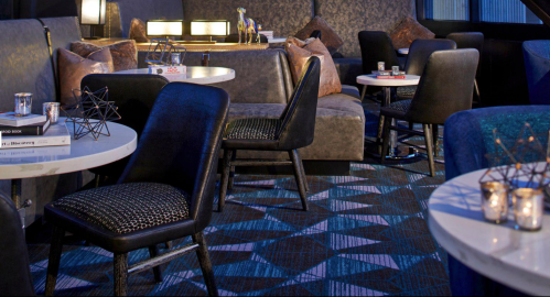 A stylish lounge area with black chairs, round tables, and decorative accents, featuring a modern geometric carpet.