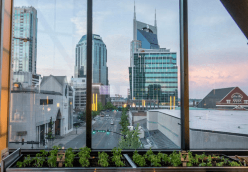 View of a city skyline with modern buildings and greenery in the foreground, captured through large glass windows.