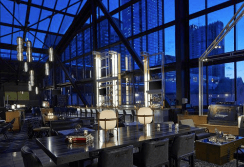 Modern restaurant interior with large windows, city skyline view, and stylish lighting at dusk.