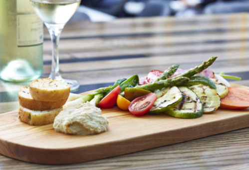 A wooden platter with grilled vegetables, hummus, sliced bread, and a glass of white wine in the background.