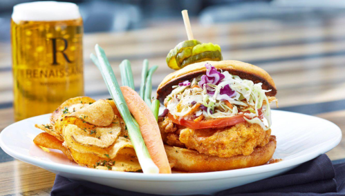 A crispy chicken sandwich topped with slaw and tomato, served with chips and a beer in the background.