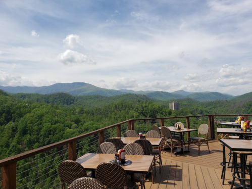 A scenic view from a deck overlooking lush green mountains and a cloudy sky, with tables and chairs set up for dining.