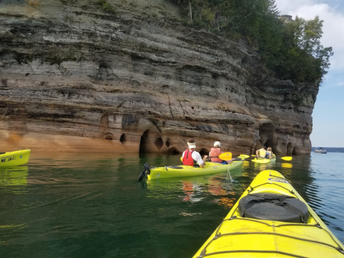 A group of kayakers paddles near rocky cliffs and caves on a calm lake. Bright yellow kayaks contrast with the scenery.