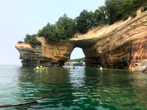 Kayakers explore a scenic rock arch surrounded by calm waters and lush greenery.