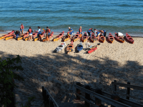 A group of people preparing kayaks on a sandy beach by the water on a sunny day.