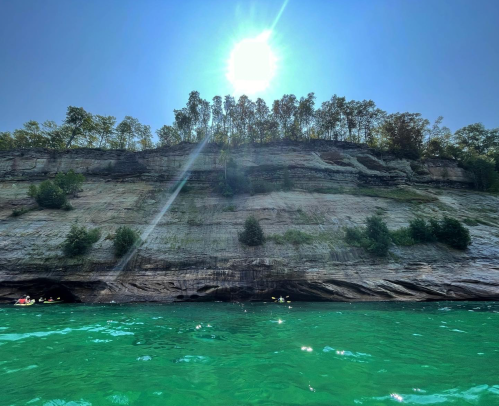 A sunlit cliff rises above vibrant green water, with trees lining the top against a clear blue sky.