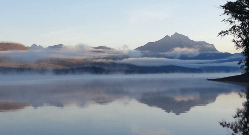 Serene lake reflecting mountains and mist under a clear sky at dawn.