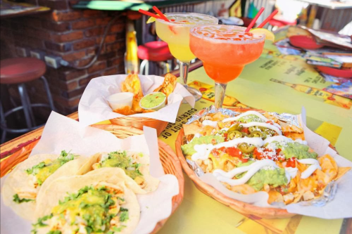 A vibrant table setting with tacos, nachos topped with guacamole, and colorful cocktails in the background.