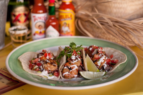 Three shrimp tacos topped with pico de gallo and lime, served on a green plate with hot sauces in the background.