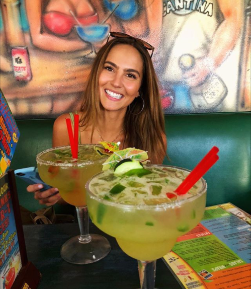 A smiling woman sits at a table with two large cocktails, surrounded by colorful decor.