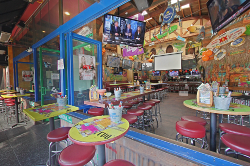 A vibrant bar interior with colorful tables, stools, and multiple TVs displaying sports, creating a lively atmosphere.