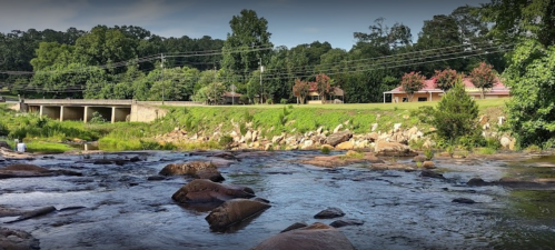 A serene river flows past a grassy bank, with trees and a few buildings in the background under a clear sky.