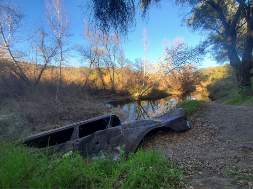 A rusted car partially submerged near a calm river, surrounded by trees and dry vegetation under a clear blue sky.