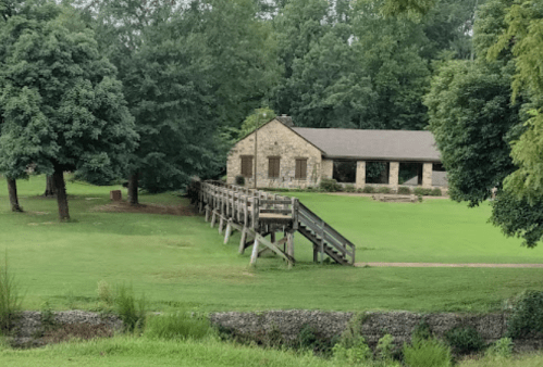 A stone building surrounded by trees and a grassy area, with a wooden walkway leading to it.