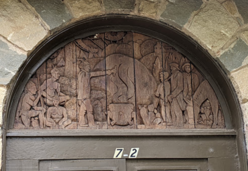 Wooden relief carving above a door, depicting various figures and a bear in a detailed, historical scene.