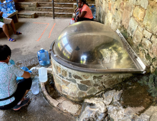 A person fills water containers at a stone water source with a dome cover, surrounded by others sitting nearby.