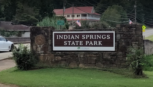 Sign for Indian Springs State Park, featuring stone structure and greenery, with a road and buildings in the background.