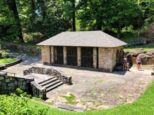 A stone building with a sloped roof surrounded by trees, with two people standing nearby on a sunny day.