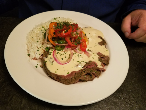 A plate of sliced beef topped with creamy sauce, served with rice and a colorful salad of peppers and onions.