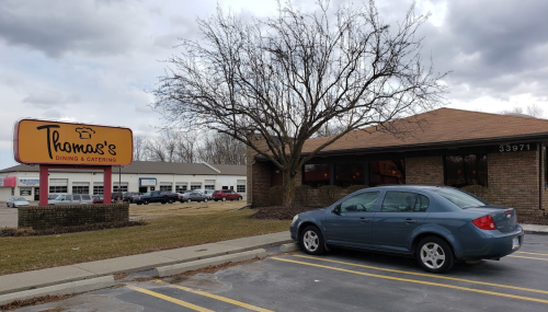 A blue car parked outside Thomas's Dining & Catering, with a sign and a cloudy sky in the background.