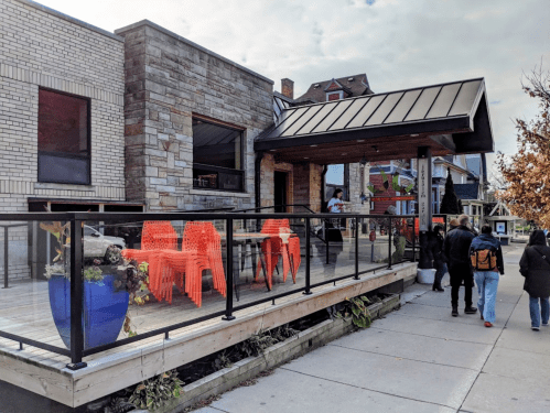 A modern building with a patio featuring orange chairs, surrounded by pedestrians on a sidewalk.