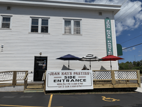 A white building with a sign for "Jean Kay's Pasties" and a side entrance, featuring outdoor seating with umbrellas.