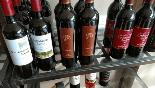 A shelf displaying several bottles of red wine with various labels, including Sycamore Lane and Terra di Dio.