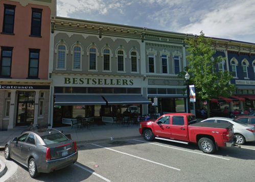 A street view of a building labeled "BESTSELLERS" with outdoor seating, flanked by other shops and parked cars.