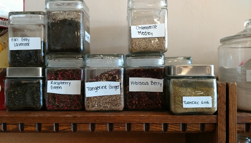 A wooden shelf displaying labeled jars of various teas and spices, including Earl Grey Lavender and Hibiscus Berry.
