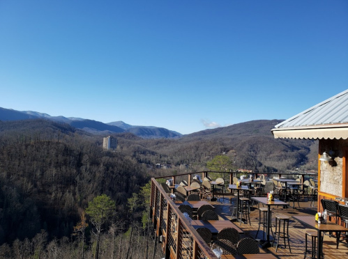 A scenic mountain view from a balcony with empty tables and chairs, under a clear blue sky.