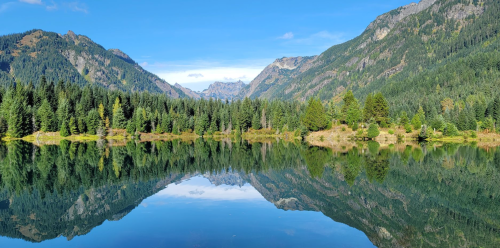 A serene lake surrounded by lush green trees and mountains, reflecting the clear blue sky and landscape.