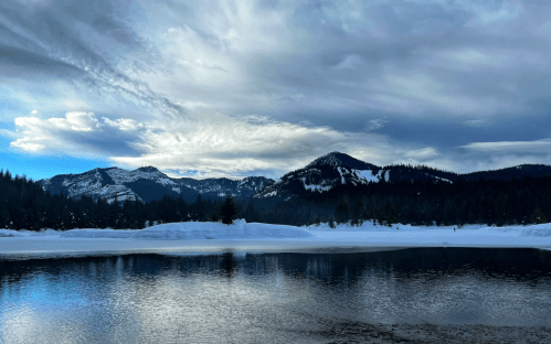 A serene winter landscape featuring a calm lake, snow-covered mountains, and a cloudy sky.