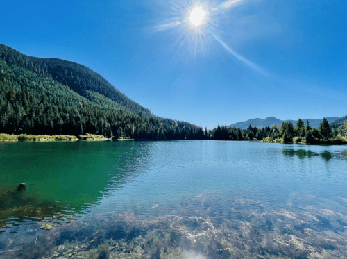 A serene lake surrounded by lush green mountains under a clear blue sky, with the sun shining brightly above.