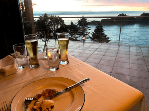 A dining table with a plate of food and two glasses of beer, overlooking a scenic ocean view at sunset.