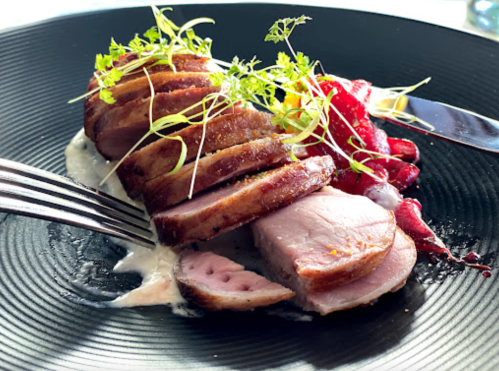 Sliced duck breast on a black plate, garnished with microgreens and accompanied by a berry sauce.