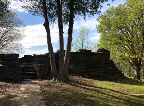 Ruins of a stone structure surrounded by trees and greenery under a blue sky with scattered clouds.