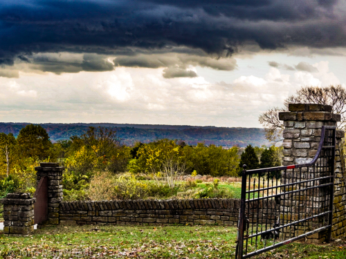 A stone gate opens to a scenic view of rolling hills under a dramatic, cloudy sky.