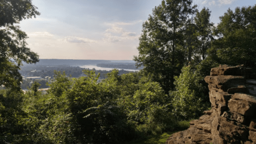 A scenic view from a rocky outcrop, overlooking a river and lush greenery under a partly cloudy sky.