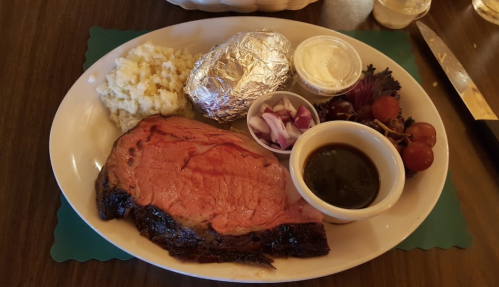 A plate with prime rib, baked potato, mashed potatoes, salad, and dipping sauce.