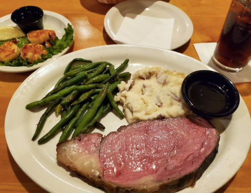 A plate with prime rib, mashed potatoes, green beans, and a side of dipping sauce, with a small plate of shrimp in the background.