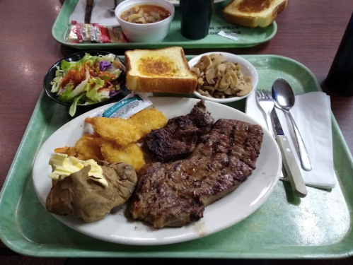 A plate with grilled steak, fish fillet, baked potato, salad, and toast, served on a green tray with utensils.