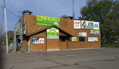 A wooden building with signs advertising food specials, drinks, and a burger named "Fort Burger." Trees in the background.
