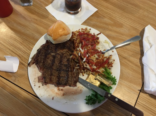 A plate with a grilled steak, a dinner roll, seasoned fries, and a side of lettuce on a wooden table.